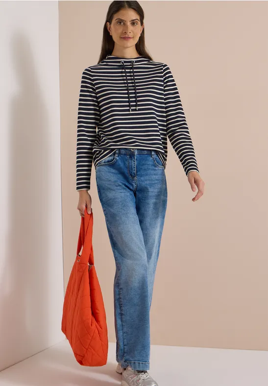 Frau in gestreiftem Pullover, weiten Jeans und orangefarbener Tasche vor heller Wand
