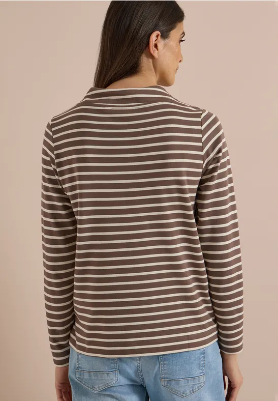 Femme de dos portant un haut à manches longues marron à rayures blanches et un jean bleu sur fond clair.