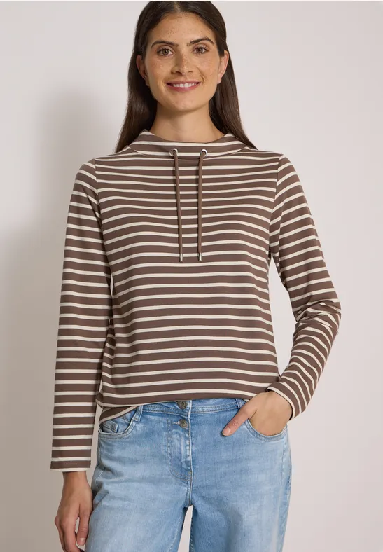 Jeune femme souriante en sweat à capuche marron et blanc à rayures, jeans clairs, une main dans la poche.