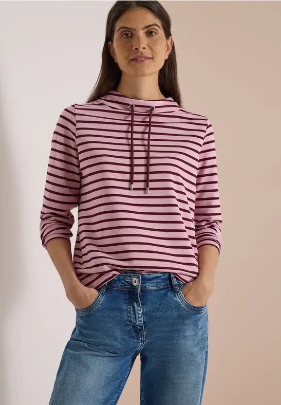 Frau in rosa-schwarz gestreiftem Kapuzenpullover und blauen Jeans vor neutralem Hintergrund.