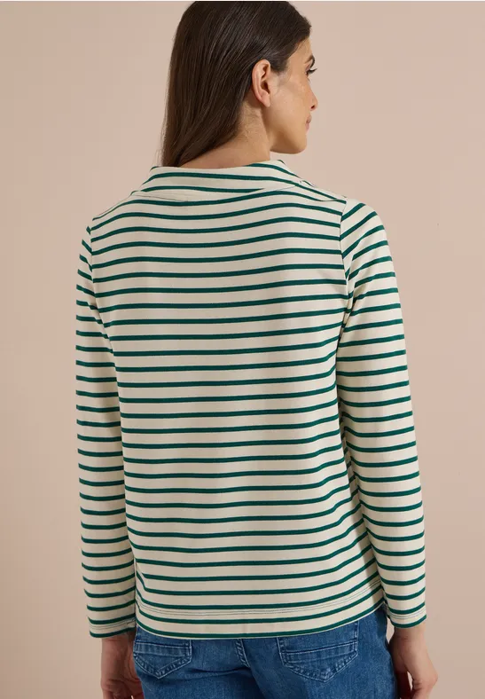 Frau von hinten in grün-weiß gestreiftem Langarmshirt und Jeans vor beiger Wand.