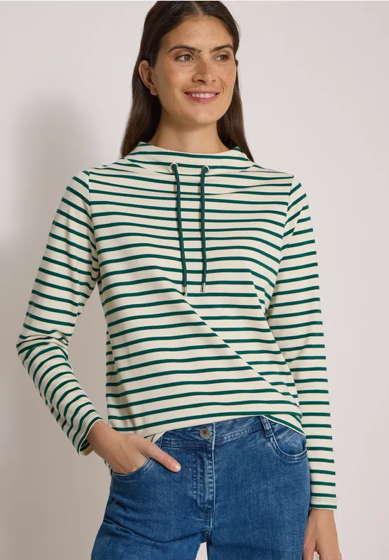 Frau lächelt in grün-weiß gestreiftem Longsleeve mit Kapuze und Jeans vor hellem Hintergrund.