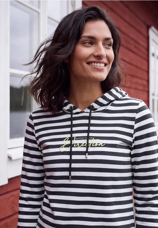 Frau im schwarz-weißen Hoodie mit gelbem Aufdruck lächelt vor roter Fassade.