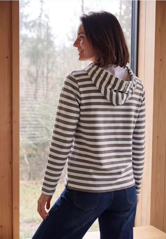 Vrouw in gestreepte capuchontrui staat bij het raam en kijkt naar het bos.