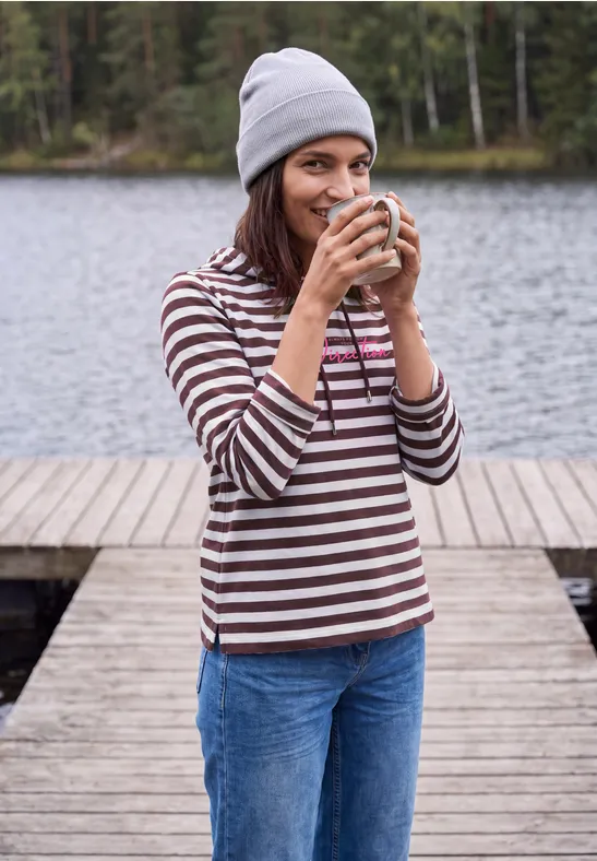 Junge Frau in gestreiftem Pullover steht auf einem Steg am See und trinkt lächelnd aus einer Tasse.