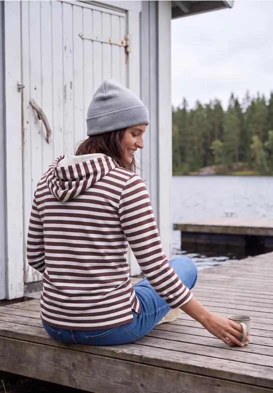 Person in gestreiftem Hoodie und Mütze sitzt auf Holzsteg am See, hält eine Tasse, Wald im Hintergrund.