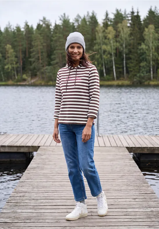 Eine lächelnde Frau in gestreiftem Pullover, Jeans und Mütze steht auf einem Steg vor einem See und Wald.