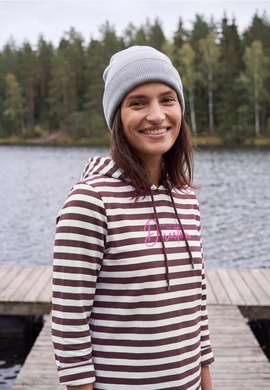 Eine lächelnde Frau in gestreiftem Hoodie und Mütze steht an einem See vor einem Wald auf einem Steg.
