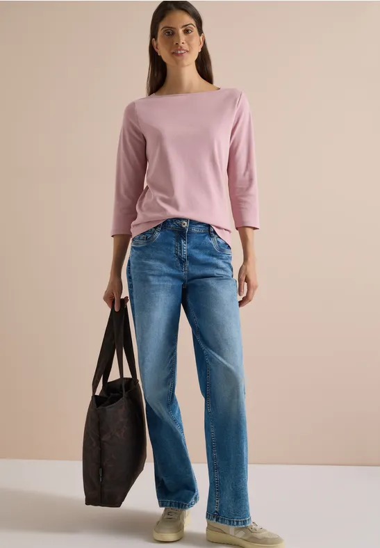 Eine Frau in rosa Dreiviertelarmshirt, blauen Jeans hält eine große, dunkle Tasche vor hellem Hintergrund.