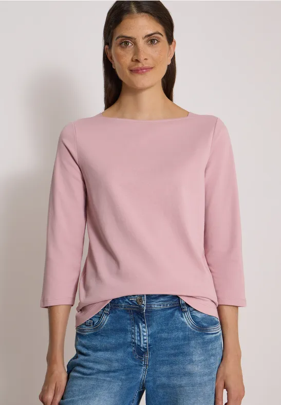 Frau trägt rosa Pullover und blaue Jeans vor neutralem Hintergrund.