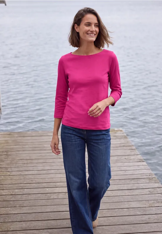 Frau in pinkem Shirt und Jeans spaziert auf einem Holzsteg am Wasser, lächelnd.