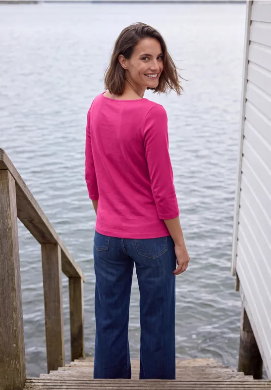 Eine lächelnde Frau in pinkem Pullover und Jeans steht auf Holztreppe am Wasser und blickt zurück.