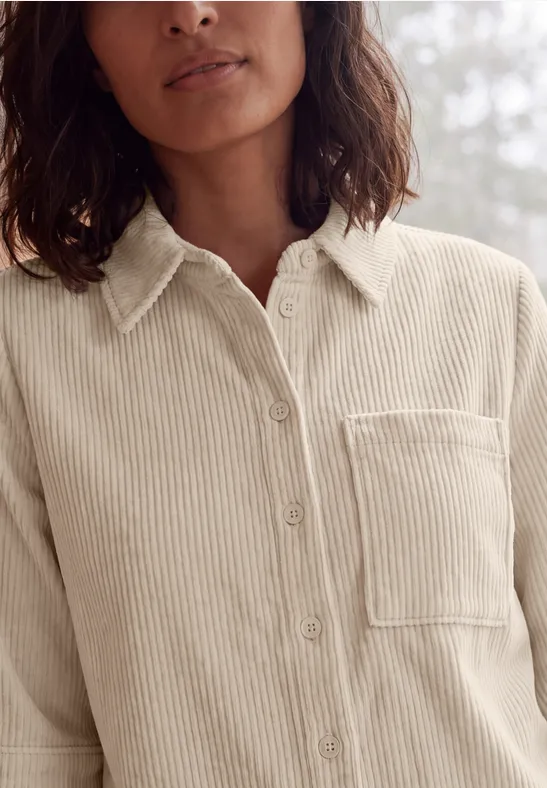 Femme porte une chemise en velours côtelé beige clair avec poche poitrine et boutons.