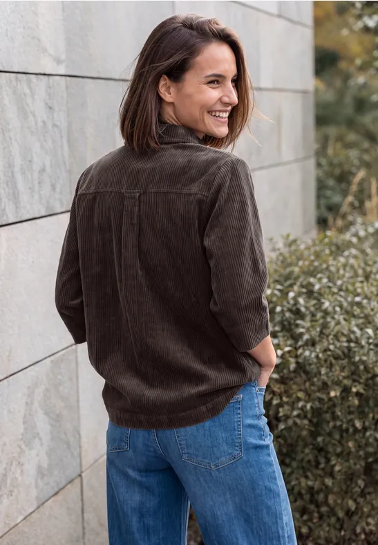 Eine Frau in brauner Cordjacke und blauer Jeans lehnt lächelnd an einer Steinmauer im Freien.
