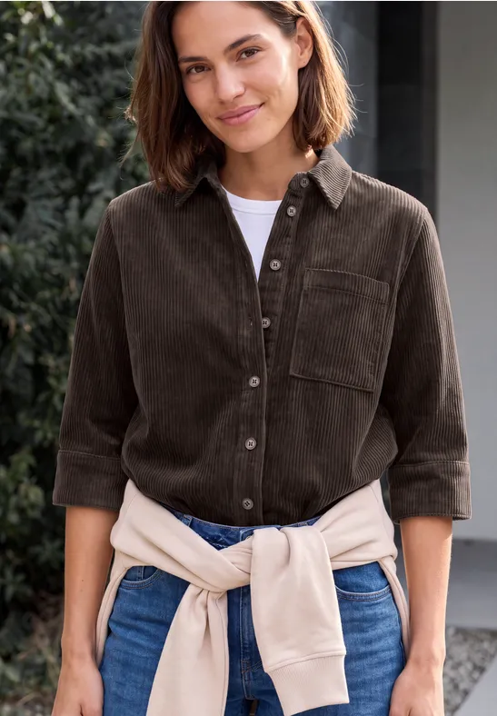 Lächelnde Frau in braunem Cordhemd, Jeans und hellbeiger Strickjacke um die Taille im Freien.