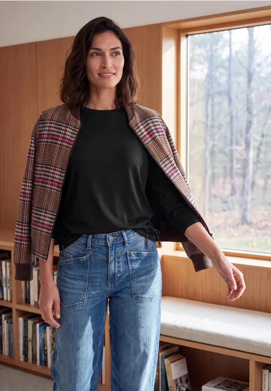 Vrouw met geruite jas over zwart shirt en jeans bij een raam in een houten interieur.
