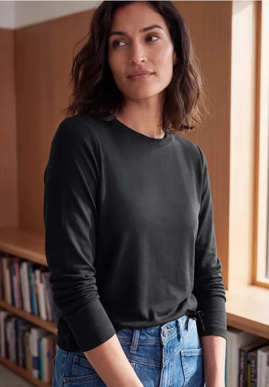 Een vrouw in een zwart shirt met lange mouwen en spijkerbroek staat nonchalant voor een boekenkast.