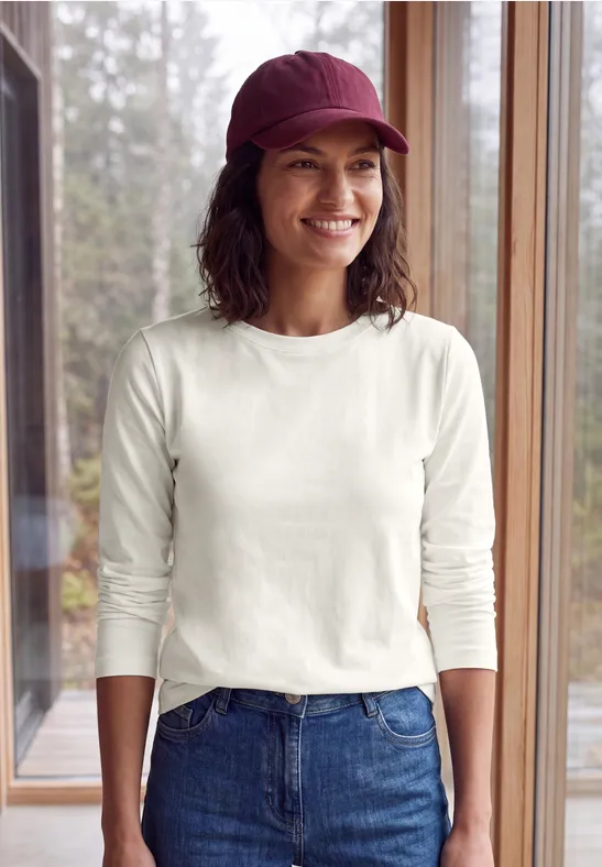 Frau in weißem Langarmshirt, Jeans und roter Mütze steht lächelnd vor großen Fenstern mit Waldblick.