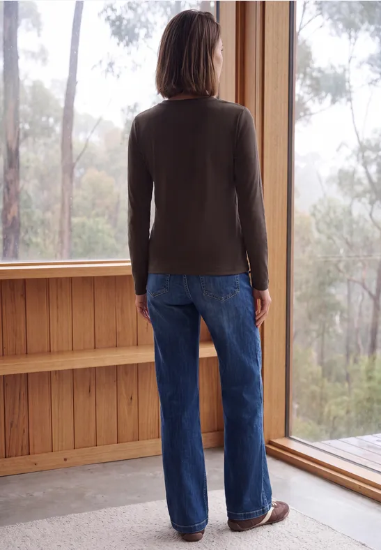 Eine Person steht in blauer Jeans und grauem Pullover vor einem großen Fenster und blickt in den Wald.