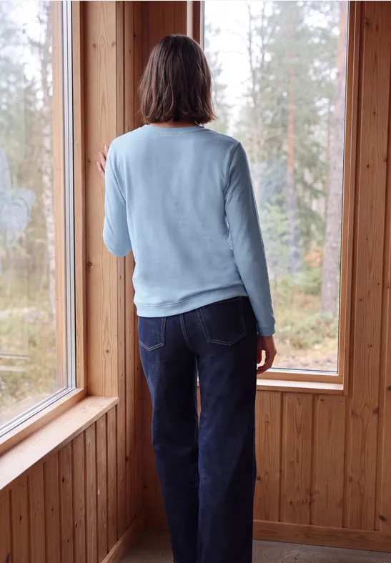 Eine Person steht in einem Holzraum und blickt durch große Fenster auf einen Wald.