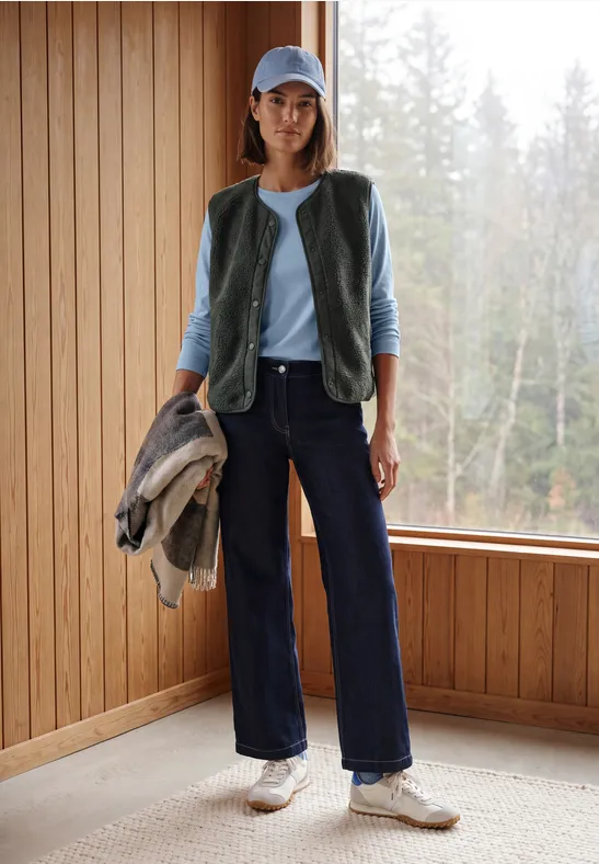 Frau im Casual-Look mit Weste, Cap, Jacke in der Hand, weiten Jeans und Sneakern in einem Holzraum vor Wald.