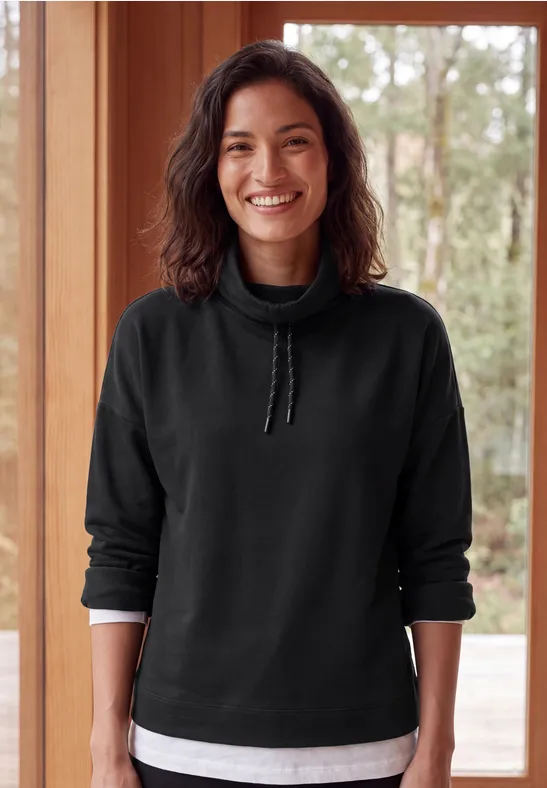 Een lachende vrouw in een zwart sweatshirt en wit T-shirt staat voor een houten raam.