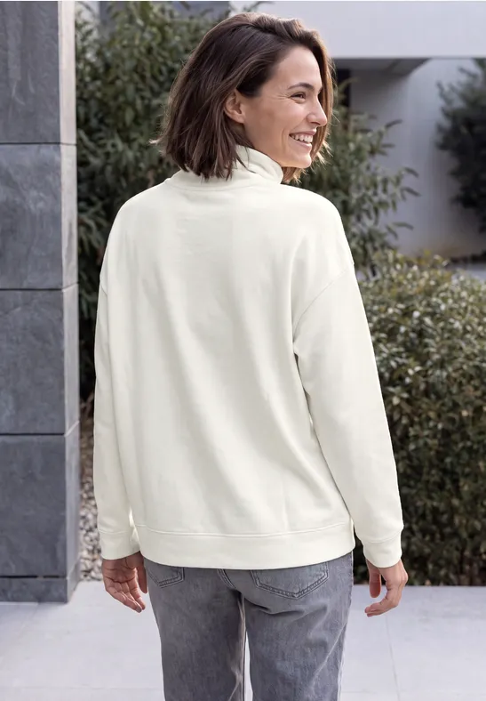 Vrouw in crème fleecevest en grijze jeans, glimlachend van opzij kijkend buiten.