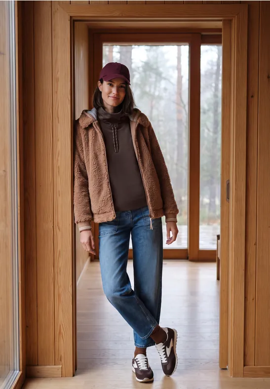 Frau in Jeans, brauner Jacke, Hoodie und Mütze steht lässig in einem hellen Holzhausflur.