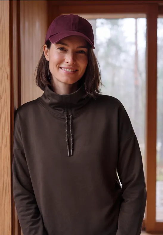 Frau mit roter Kappe und dunklem Pullover lächelt vor hellem Fenster in Holzinterieur.