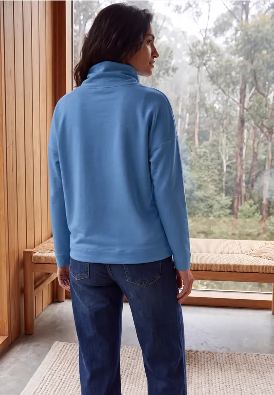 Frau in blauer Fleecejacke und Jeans steht auf Holzveranda, schaut in nebligen Wald.