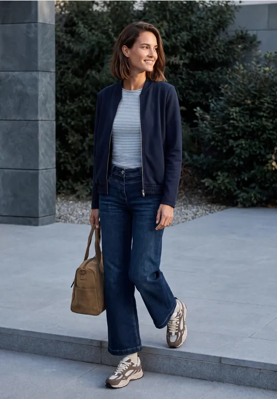 Eine Frau in Jeans, blauem Cardigan und Sneakern spaziert vor modernem Gebäude und trägt eine Beige-Handtasche.