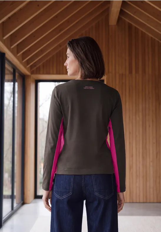Frau steht in Holzbau, trägt olivgrünen Pullover mit pinken Seiteneinsätzen und Jeans.