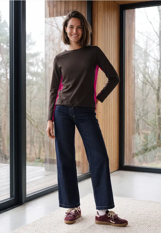 Eine lächelnde Frau in modernem Longsleeve und weiten Jeans steht vor großen Fenstern in einem hellen Raum.
