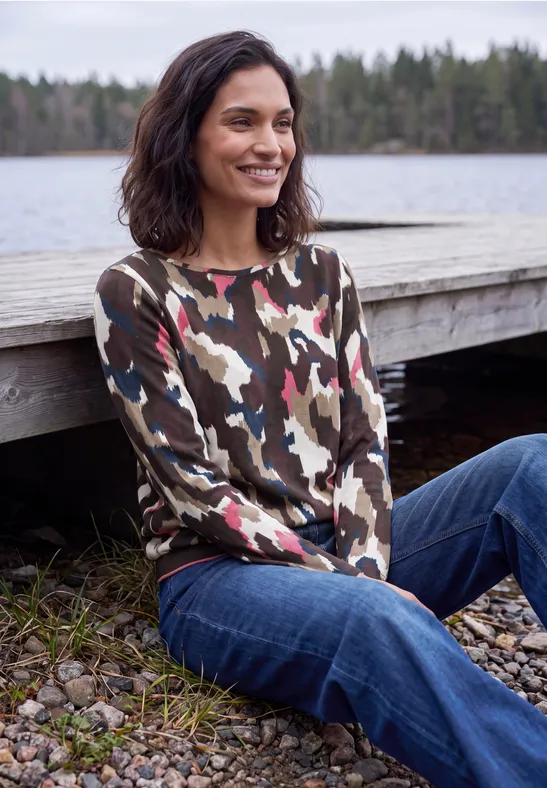 Eine Frau sitzt lächelnd am Holzsteg an einem See, trägt Camouflage-Pullover und Jeans.