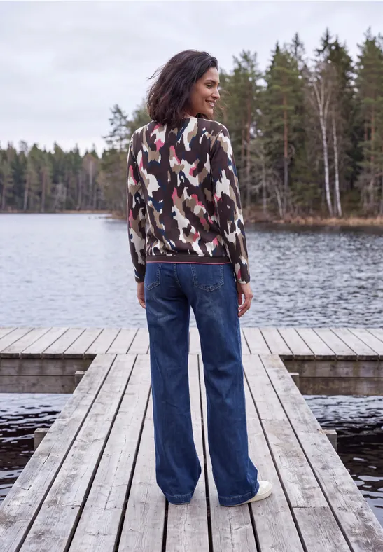 Frau steht im Camouflage-Pullover und weiter Jeans mit dem Rücken zur Kamera auf einem Steg am See.