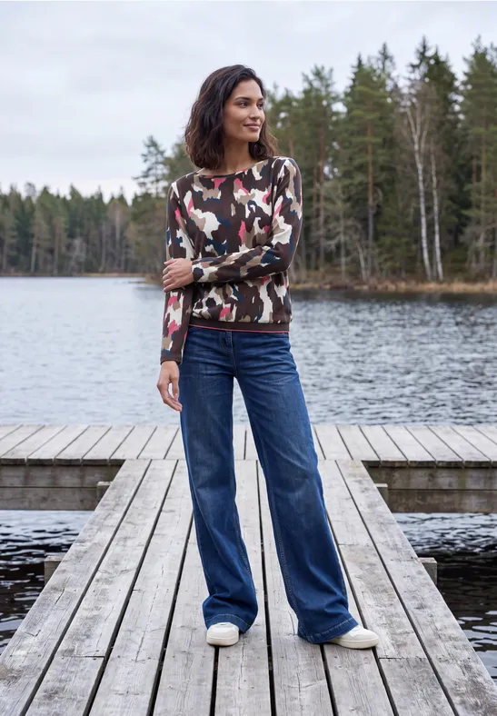 Eine Frau in Camouflage-Pullover und weiten Jeans steht auf einem Holzsteg an einem Waldsee.