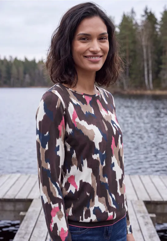 Eine Frau im bunten Pullover steht an einem Steg vor einem ruhigen Waldsee.