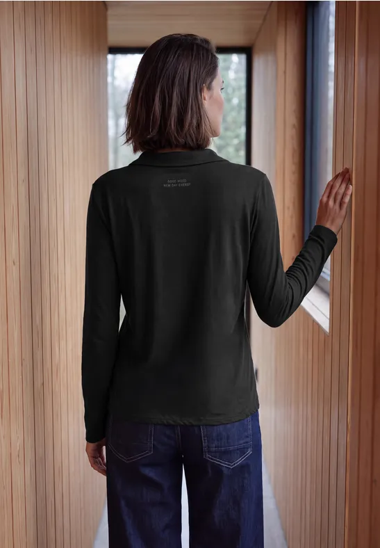 Person von hinten in schwarzem Langarmshirt und Jeans, steht im hellen Holzflur am Fenster.