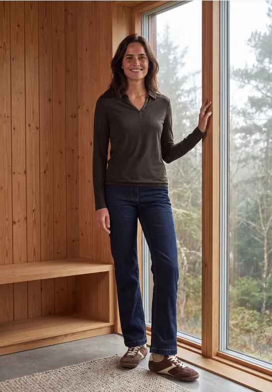 Frau in lässiger Kleidung (Pullover, Jeans, braune Schuhe) steht in modernem Holzinnenraum am Fenster.