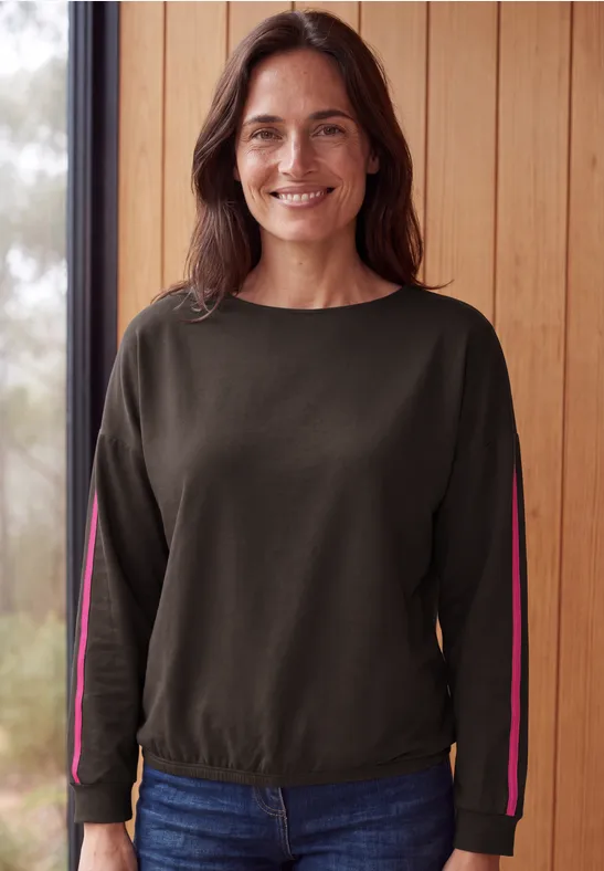 Eine lächelnde Frau in grünem Sweatshirt und Jeans vor einer Holzwand.