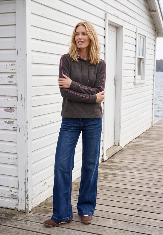 Een glimlachende vrouw in donkere trui en wijde jeans staat voor een witte houten wand op een veranda.