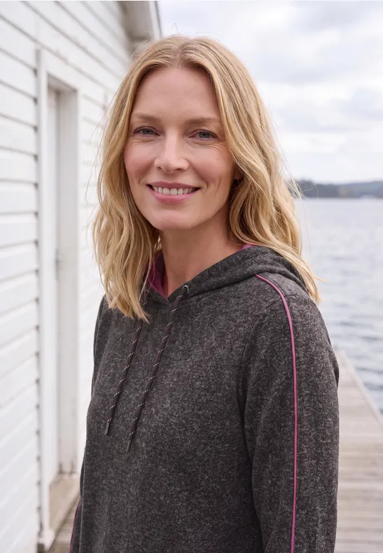 Een glimlachende blondine in een grijze hoodie voor een wit boothuis aan het water.