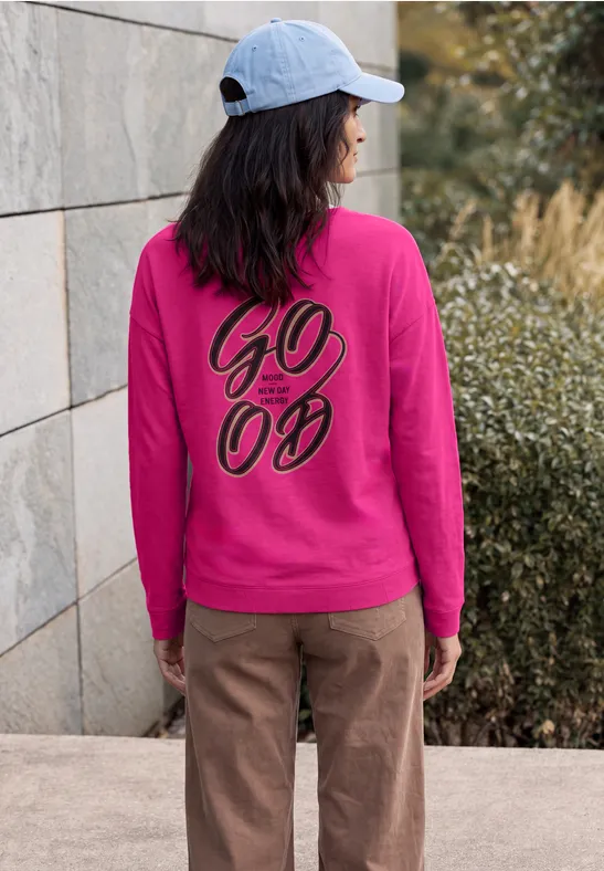Vrouw in roze sweater met zwarte 'GO 00'-print op de rug en lichtblauwe pet, staand voor een muur.