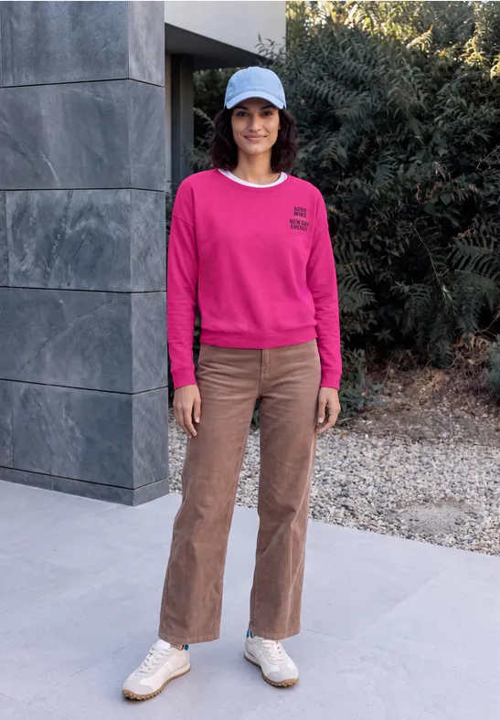 Vrouw in roze sweater, bruine broek, witte sneakers en pet voor een modern huis met struiken.
