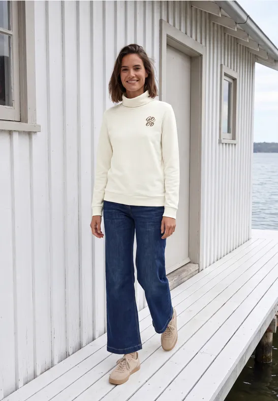 Vrouw in lichte trui en wijde jeans loopt glimlachend over een steiger langs een wit huis aan het water.