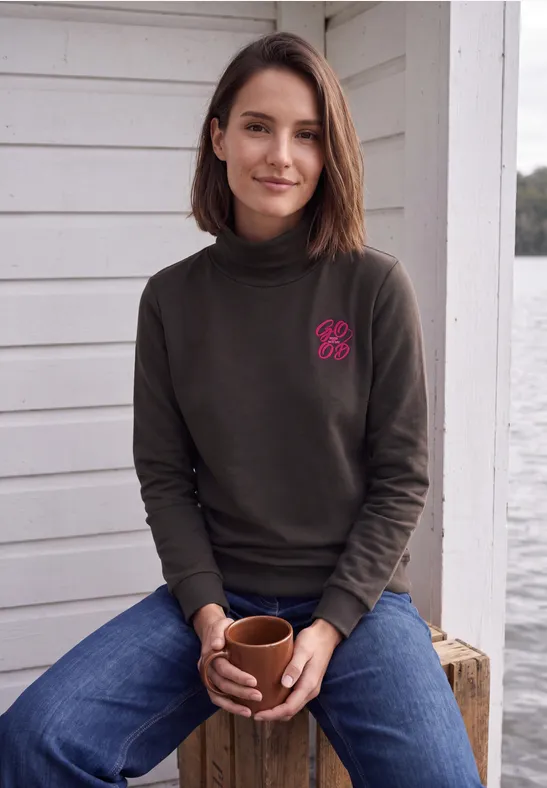 Een vrouw zit ontspannen met een kop koffie, draagt coltrui en spijkerbroek voor een wit huis
