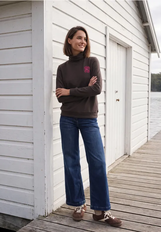 Een vrouw staat ontspannen bij een wit strandhuis op de pier, met trui, spijkerbroek en bootschoenen.