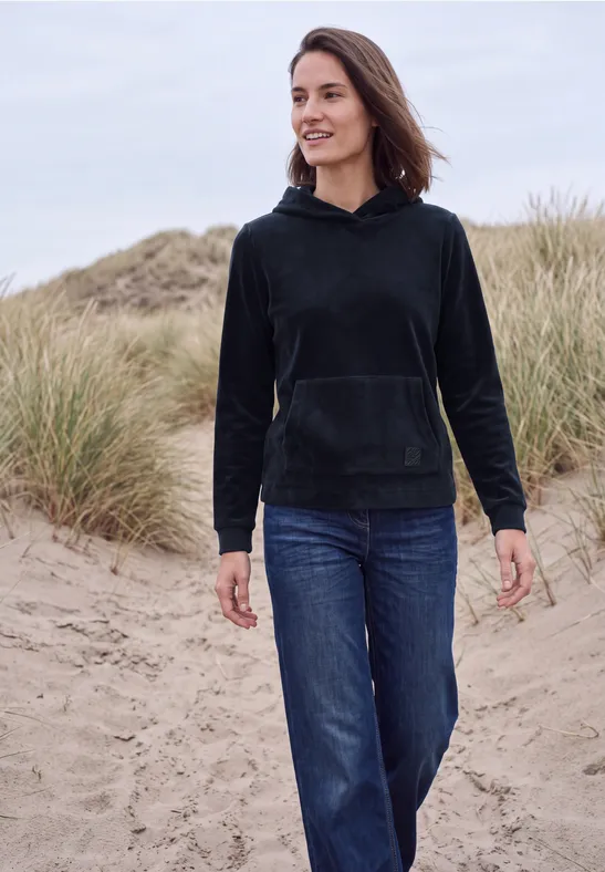 Eine Frau in schwarzer Kapuzenjacke und Jeans spaziert am Strand zwischen Dünen.