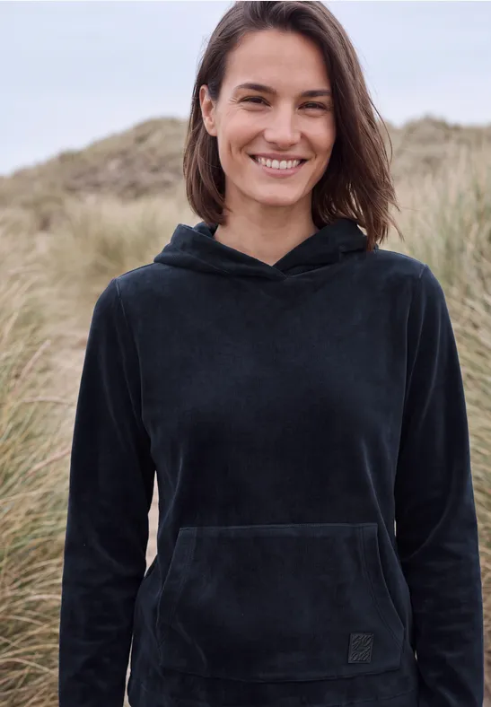 Lächelnde Frau in schwarzem Kapuzenpullover vor Dünen und Strandgras.