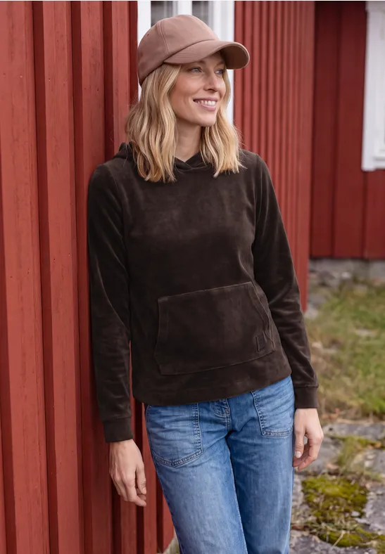 Eine Frau in dunkelbraunem Hoodie, Jeans und Cap lehnt lächelnd an einer roten Holzwand.